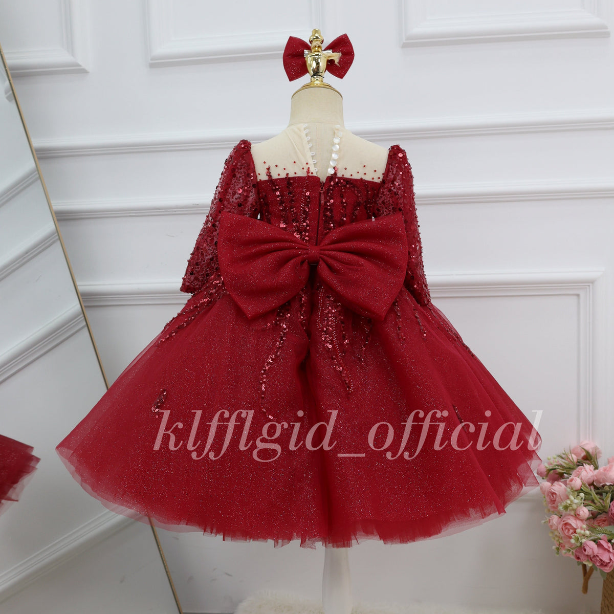 Back view of a burgundy girls' party dress with a large satin-look bow, sparkling long sleeves, a sheer upper back, and a voluminous glittering skirt.