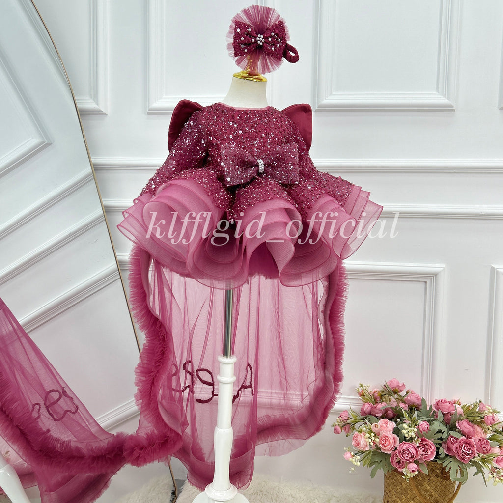 Close front view of a burgundy sequin tulle princess dress with a glittering bodice, oversized waist bow, sculpted layered skirt, and floor-length sheer tulle panels.