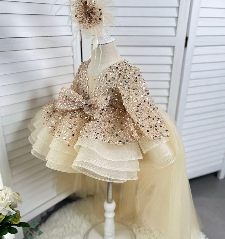 Side view of a champagne girls' princess gown featuring a glittering sequin bodice, long sleeves, a large waist bow, layered ivory ruffles, and a long tulle train.