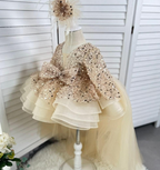 Side view of a champagne girls' princess gown featuring a glittering sequin bodice, long sleeves, a large waist bow, layered ivory ruffles, and a long tulle train.