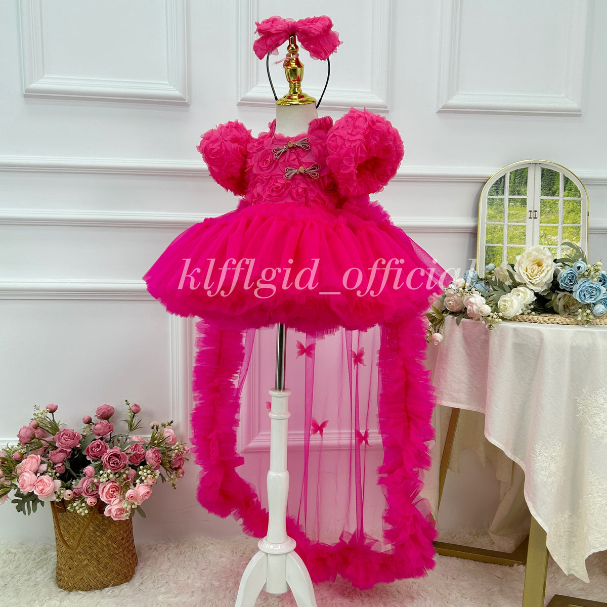 Three-quarter front view of a vibrant hot pink floral princess dress with textured rose details, rounded puff sleeves, decorative front closures, a full skirt, and an extended sheer train.
