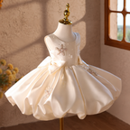 Three-quarter view of an ivory satin-look flower girl gown featuring a structured bodice, decorative butterfly appliques, floral details, a statement bow, and a voluminous skirt.