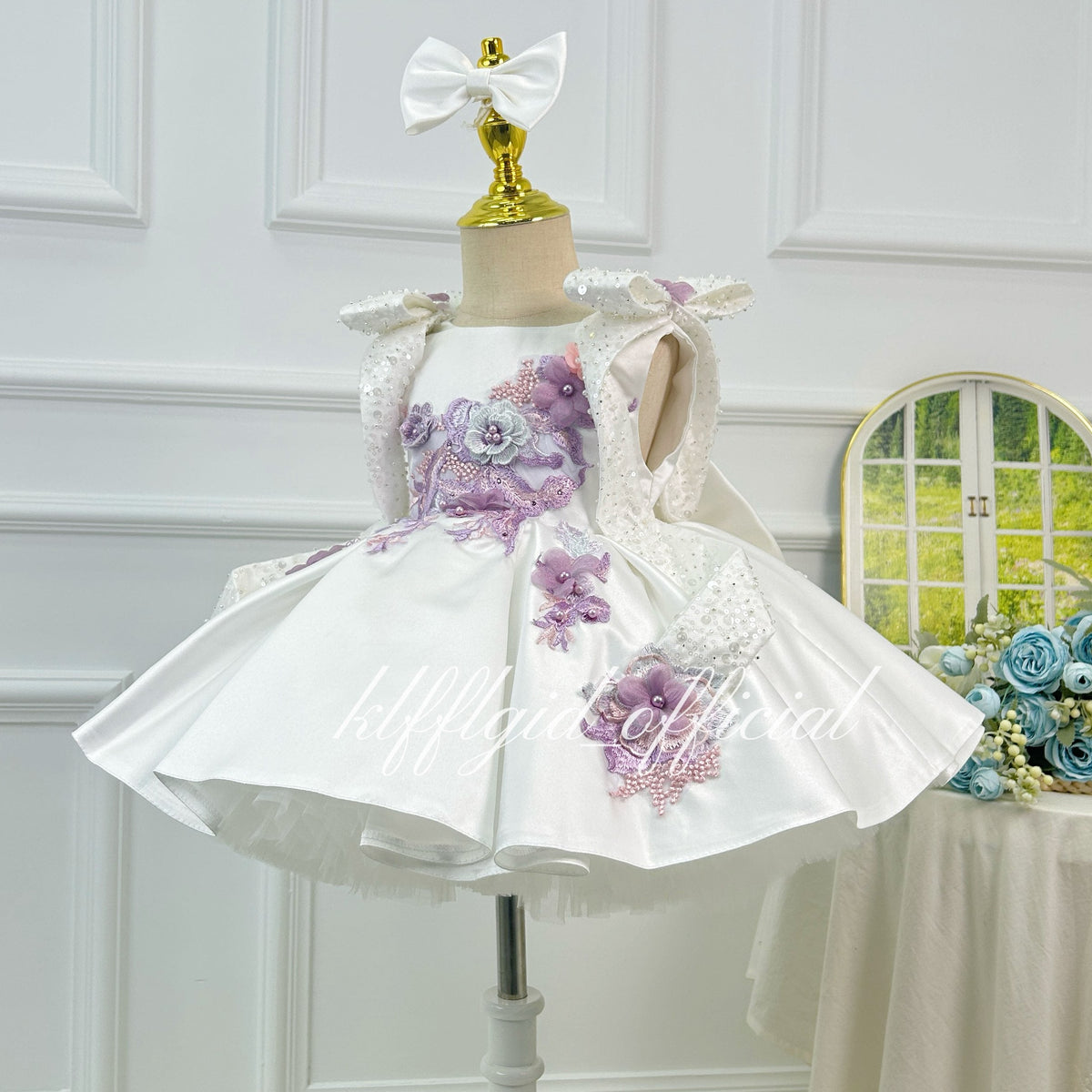 Angled view of an ivory girls' birthday dress featuring pink and purple floral embroidery, 3D flowers, decorative shoulder straps, and a structured flared skirt.