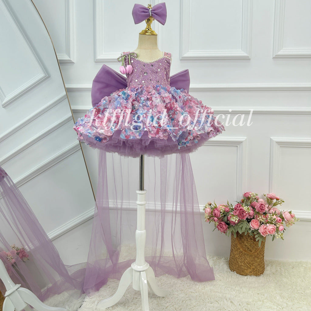 Front view of a lavender girls' princess gown featuring a sparkling bodice, pink and blue floral skirt decorations, an oversized purple bow, and a long sheer tulle train.
