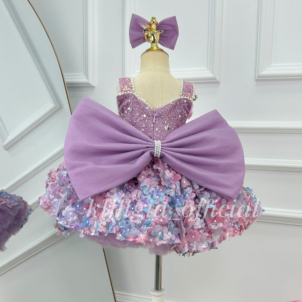 Close-up of the back of a lavender princess dress featuring an oversized purple bow with pearl-like center trim, a sparkling bodice, and colorful floral skirt decorations.