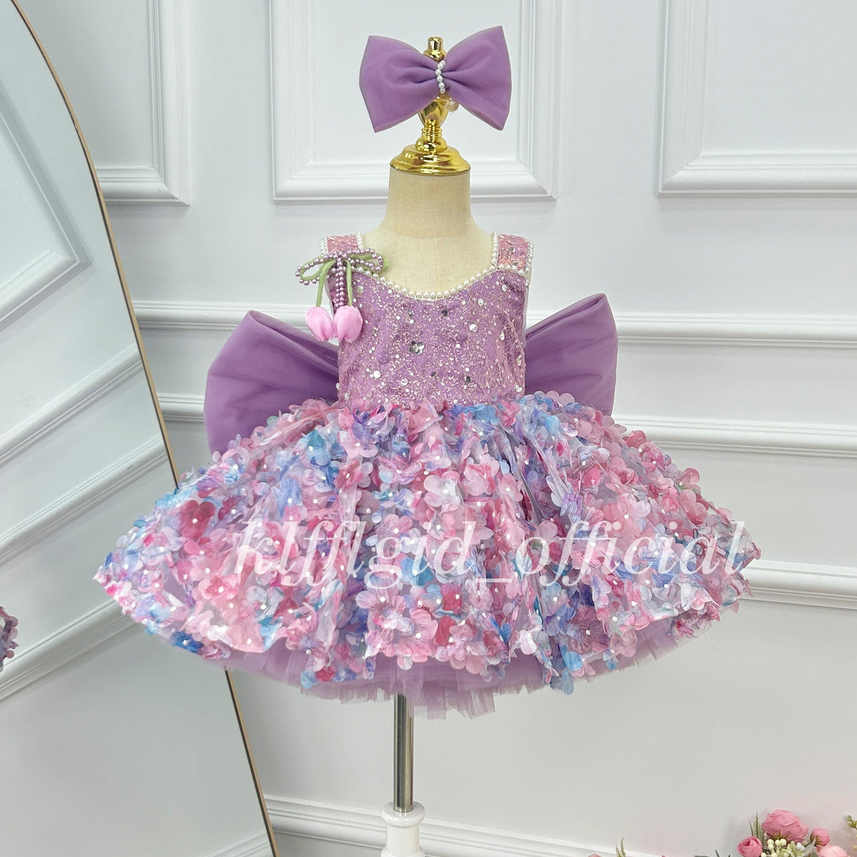 Close-up of a lavender girls' princess dress showing a sparkling embellished bodice, pink and blue floral skirt details, pearl-like neckline trim, and decorative pink flower accents.