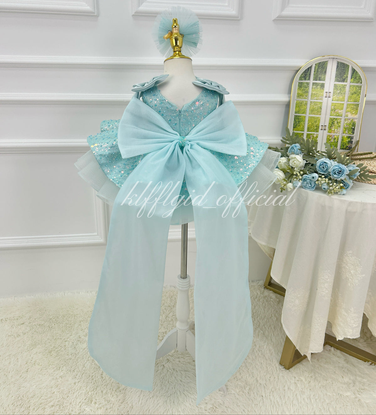 Back view of a light blue sequin princess dress for girls featuring a large matching bow, long flowing fabric tails, and a layered skirt.