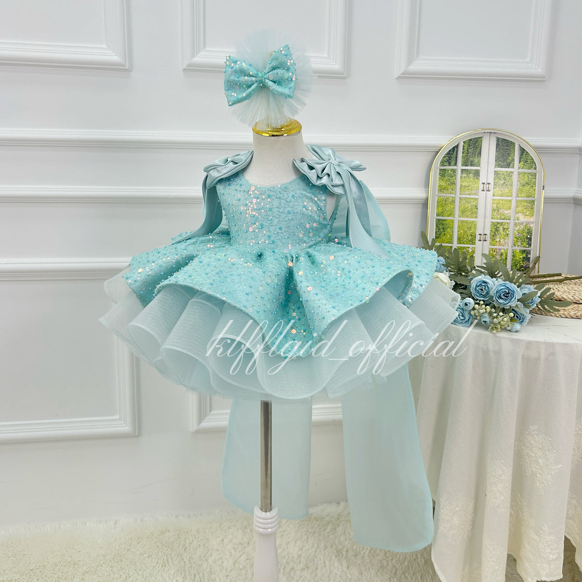 Three-quarter view of a light blue sequin tulle gown for girls with a sparkling bodice, flared layered skirt, shoulder ruffles, and long trailing fabric details.