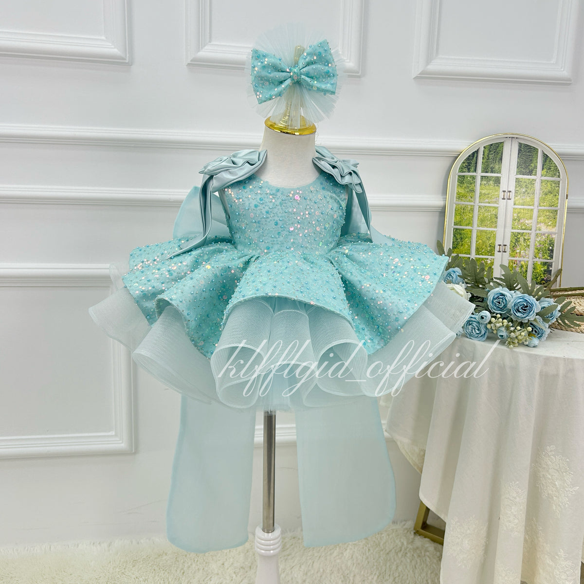 Front view of a light blue girls' sequin tulle princess dress with a sparkling bodice, layered flared skirt, shoulder ruffles, and matching bow headpiece.
