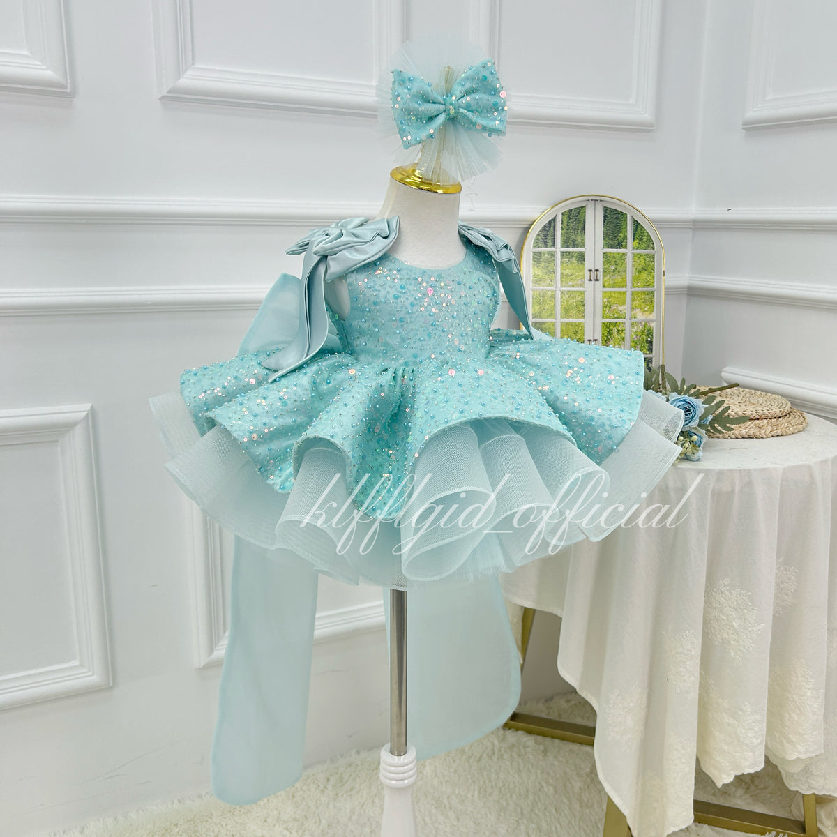 Angled view of a light blue girls' birthday dress with shimmering sequin details, a voluminous tulle skirt, ruffled shoulders, and flowing back accents.