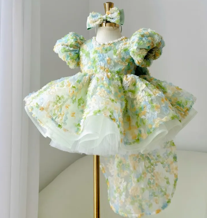 Angled view of a pastel floral baby princess gown with voluminous puff sleeves, a full flared skirt, white underskirt layers, and delicate green, blue, yellow, and ivory floral details.