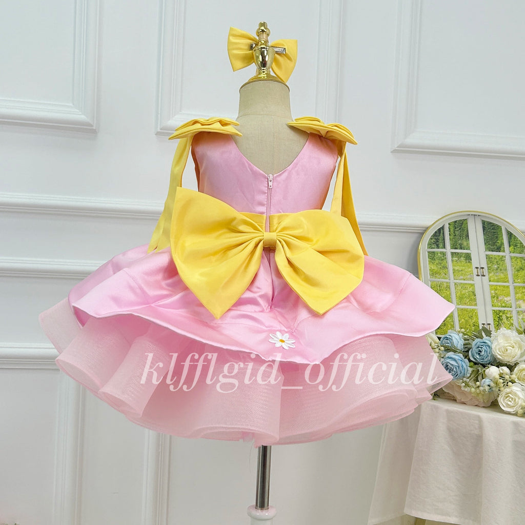 Back view of a pink girls' birthday dress featuring a large yellow bow, a back zipper, yellow shoulder details, and a full layered tulle skirt.