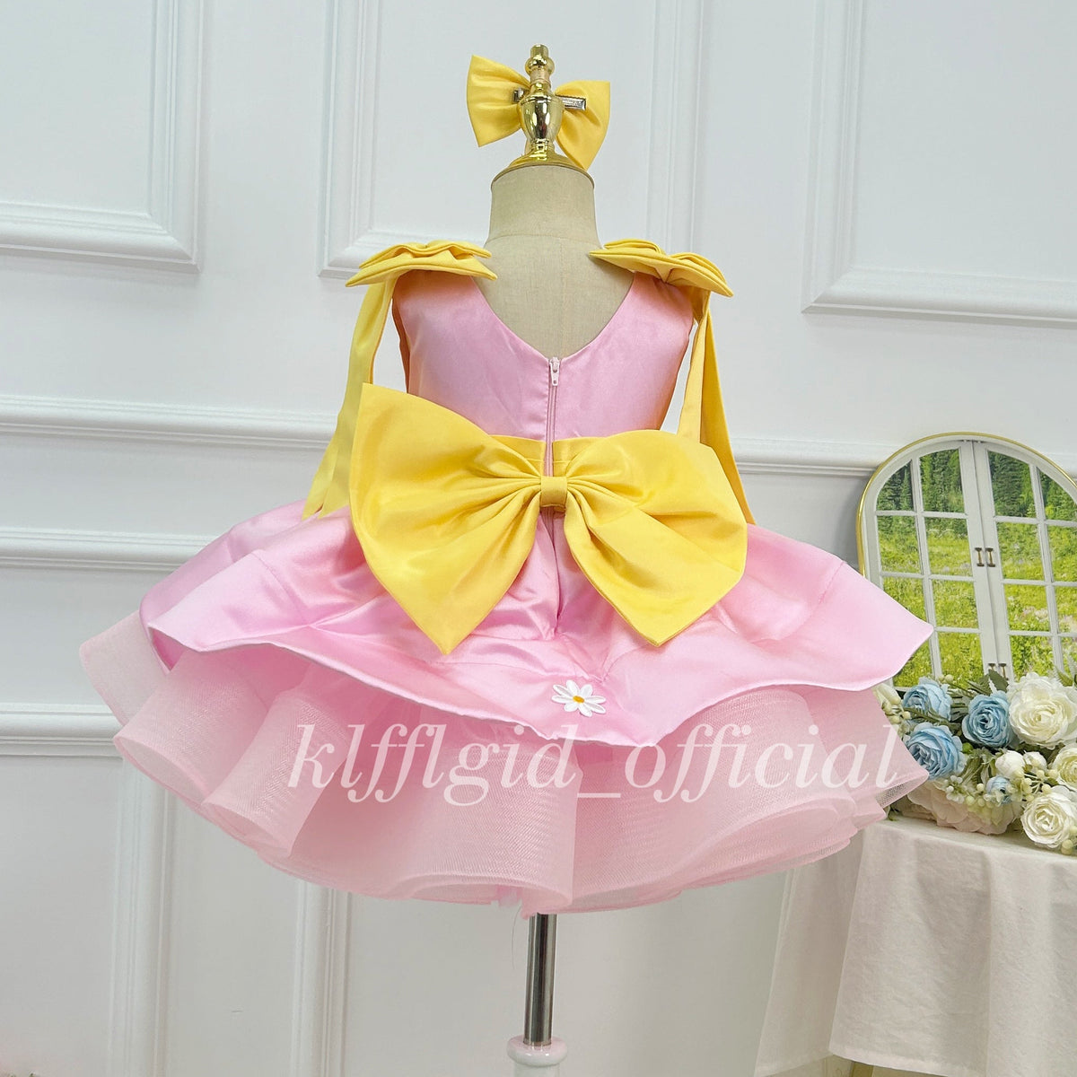 Back view of a pink girls' birthday dress featuring a large yellow bow, a back zipper, yellow shoulder details, and a full layered tulle skirt.