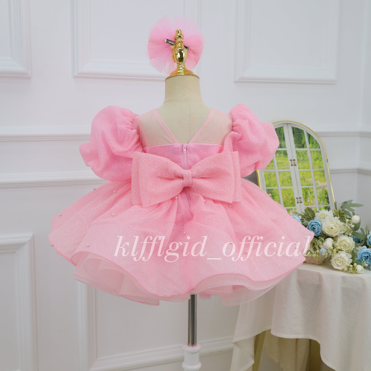 Back view of a pink girls' princess dress featuring a large glitter bow, puff sleeves, a sheer V-shaped back neckline, and a layered skirt