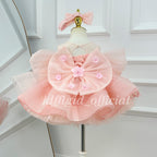 Back view of a blush pink princess dress with an oversized sheer bow decorated with small flowers, a lace-up bodice, and a layered skirt.