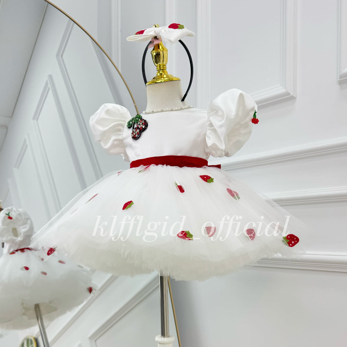 Angled view of a white strawberry-themed toddler party dress with puff sleeves, a red waistband, a decorative strawberry brooch, and a full tulle skirt with strawberry prints.