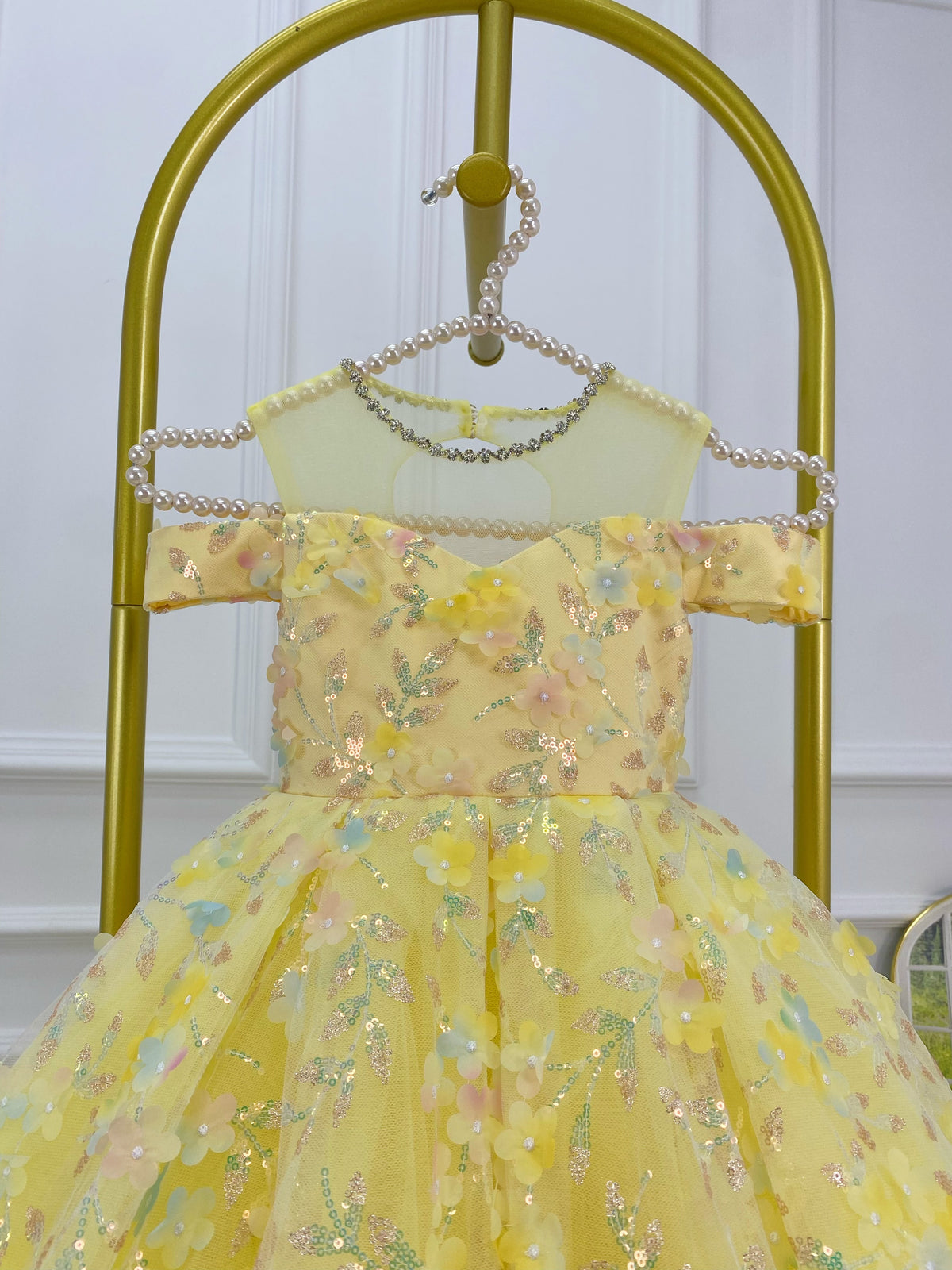 Close-up of a yellow girls' dress bodice showing an off-the-shoulder neckline, pastel floral appliqués, shimmering decorative accents, and a fitted waist.